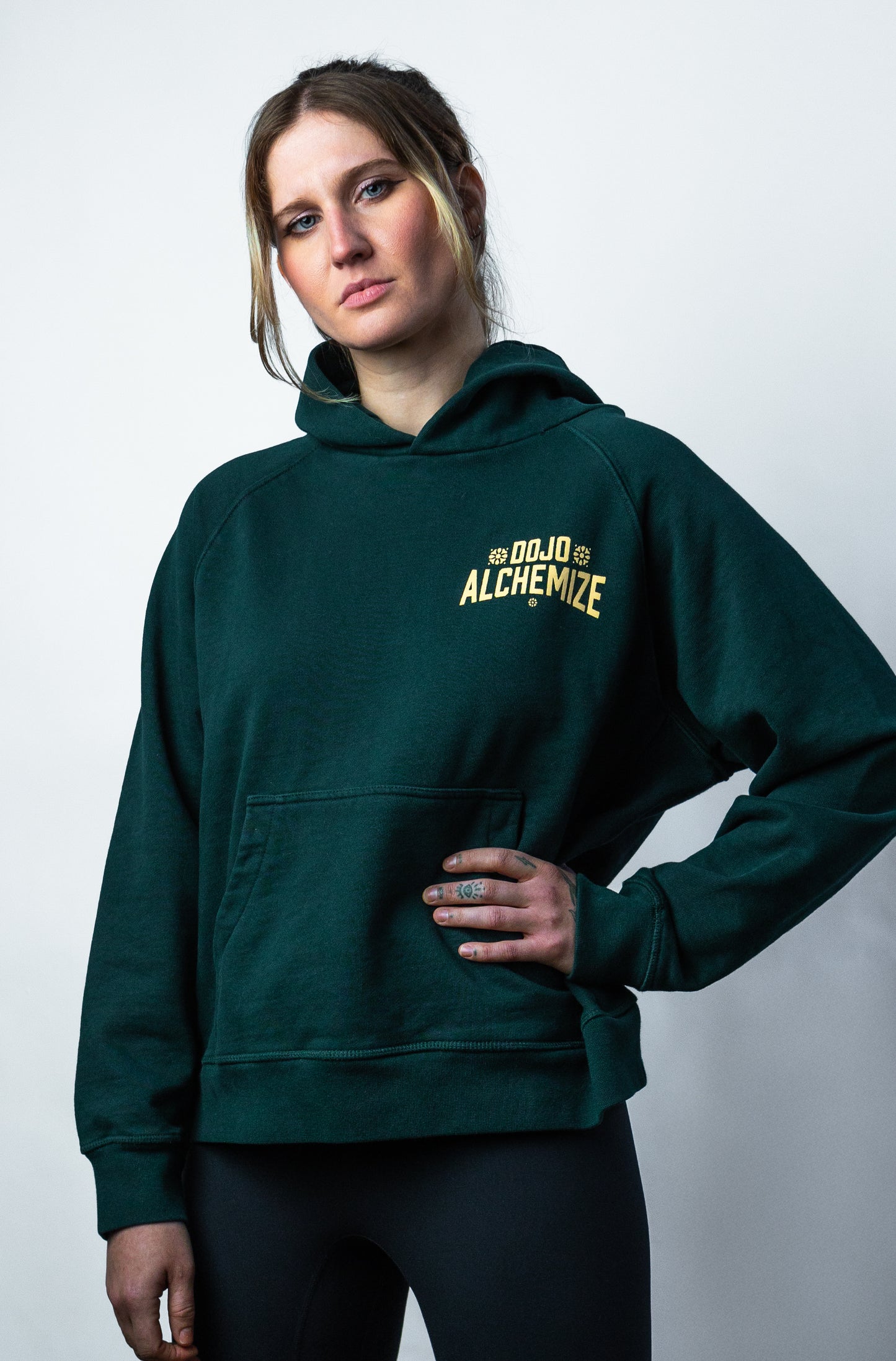 Women's forest green front pocket hoodie on model. "Dojo Alchemize" written across pocket. No drawstring.