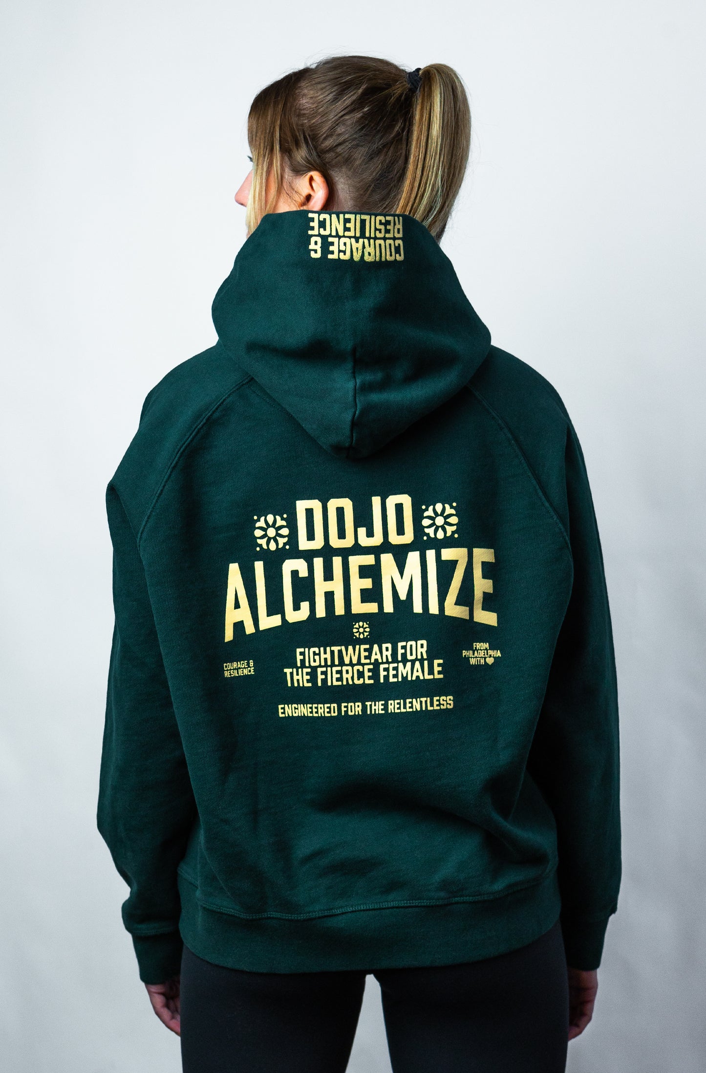 Women's forest green hoodie on model. Written across back "Dojo Alchemize Fightwear for the Fierce Female Engineered for the relentless".